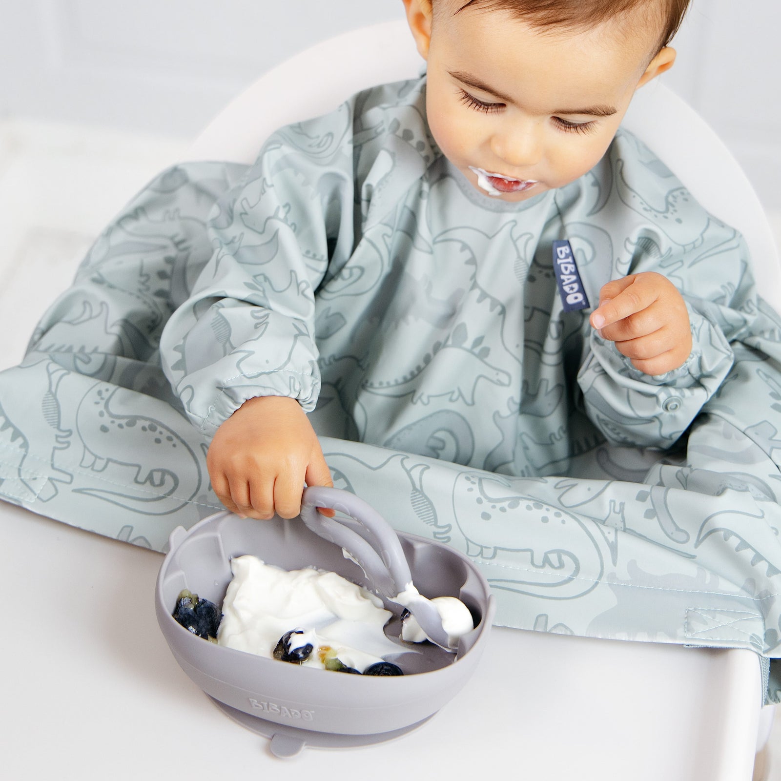 A baby wearing a light blue dinosaur-print bib sits in a white high chair, eating yogurt from a matching gray bowl with a spoon.