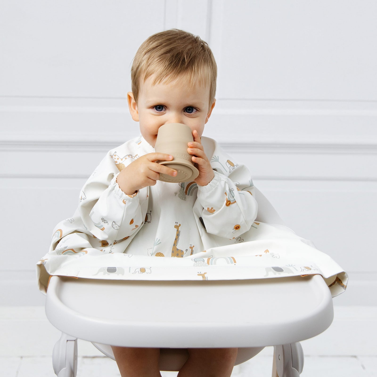A toddler wearing a cream long-sleeve shirt with a small animal print sits in a white high chair, holding a beige ceramic cup against a white paneled wall background.
